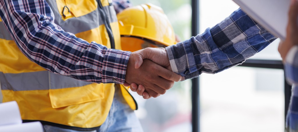 Two construction workers in plaid shirts handshake. One wears a yellow safety vest and holds a hard hat. The tone conveys agreement and professionalism.