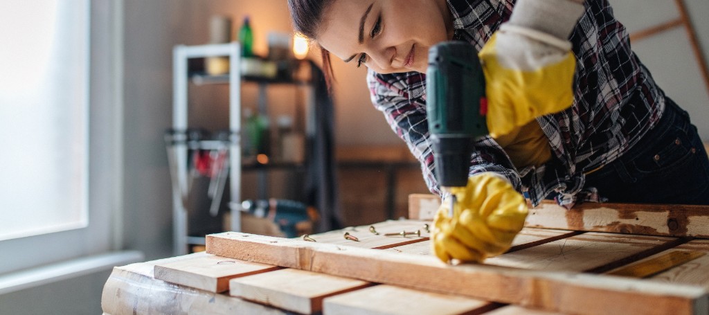 A focused child in safety goggles and yellow gloves uses a power drill to build a wooden project at a bright workshop bench with care.