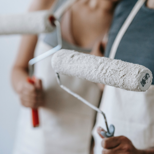 People holding paint rollers with white paint, wearing aprons. The scene suggests home renovation, conveying a collaborative and productive atmosphere.