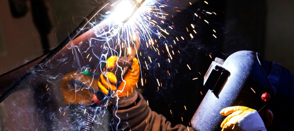 Welder in a dark workshop strikes a bright arc, sending orange sparks across a protective helmet and gloved hands; dramatic, industrious scene.
